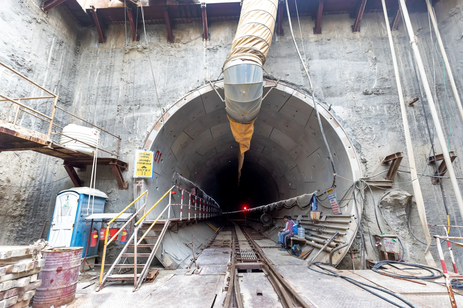 Entrance to a concrete-lined tunnel under construction, with rail tracks, a ventilation duct and access stairs