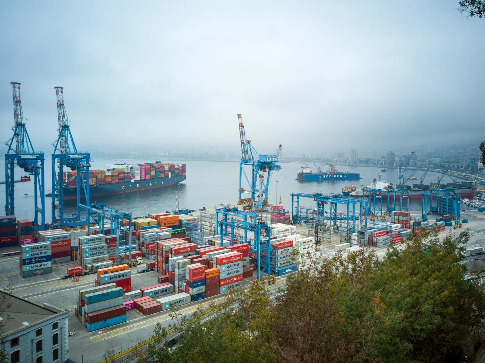 Aerial view of a hazy container port with blue cranes, stacked containers and ships